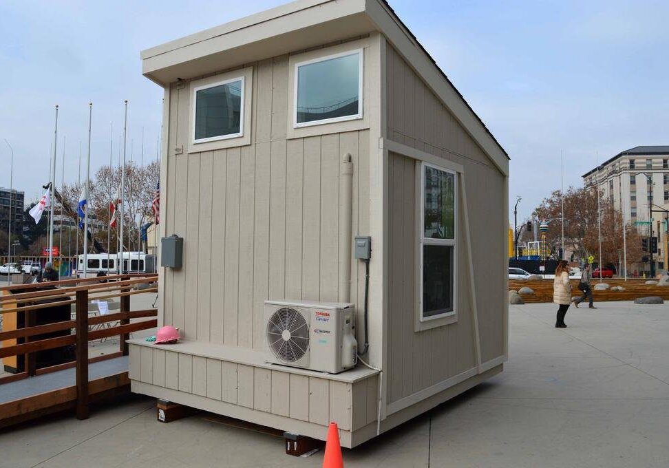 Small beige portable building with windows set on a city sidewalk, featuring an air conditioning unit on the side, surrounded by a wooden ramp and barriers. flags and buildings in the background.